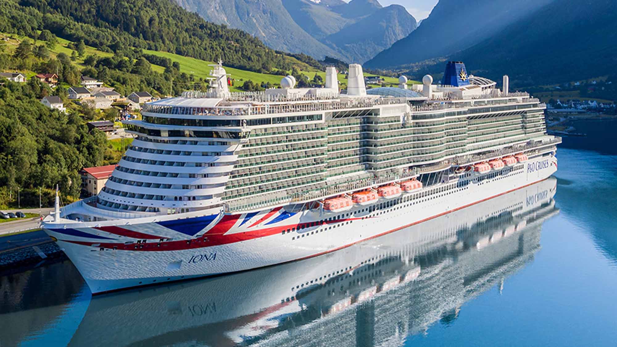 A large white cruise ship with red and blue accents, docked in calm blue water with green hills and mountains in the background. The ship is shown from an elevated angle, with its reflection visible on the water surface.